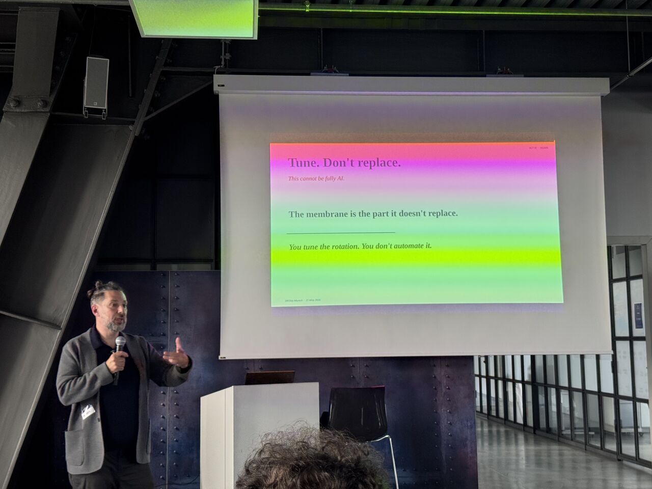 Andrea Valenti on stage at SREday Munich 2026, beside a slide reading 'Tune. Don't replace. The membrane is the part it doesn't replace.'